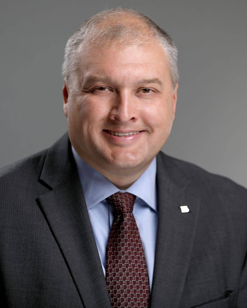 Headshot of Kevin Howell smiling, wearing a dark gray suit, blue shirt, and burgundy tie.
