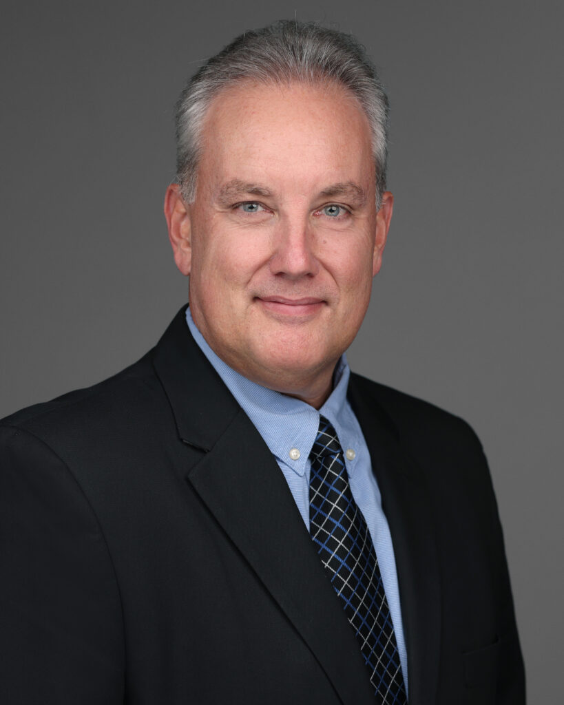 Headshot of Tom Tyra smiling and wearing a dark suit, blue shirt, and dark tie.