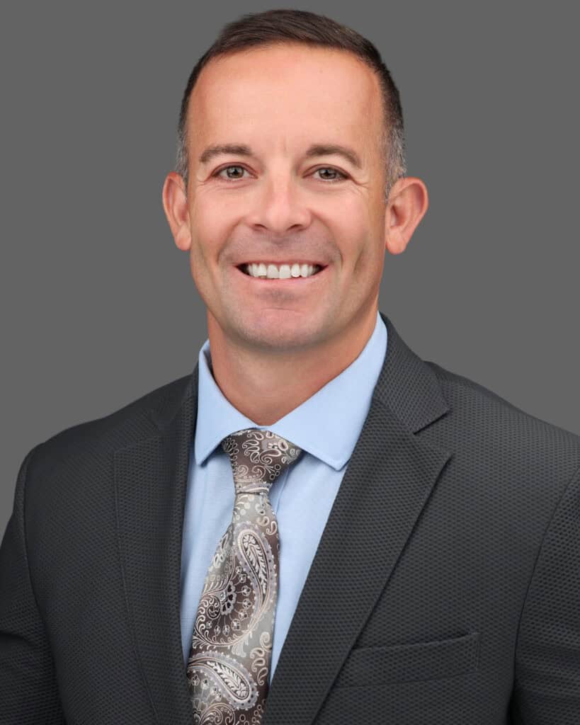 Friendly professional portrait of a smiling man in business attire against a gray background.