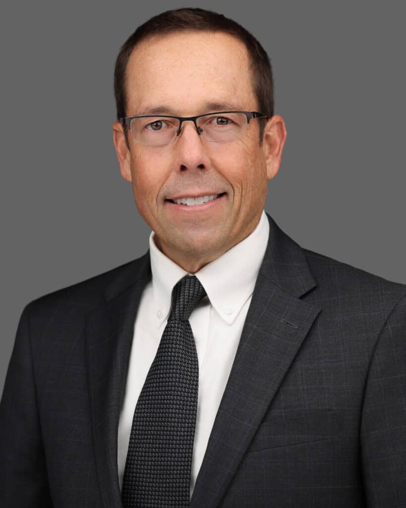 Professional headshot of Kent Bontrager wearing a dark suit, white dress shirt, and a patterned dark grey tie, posed against a neutral background.