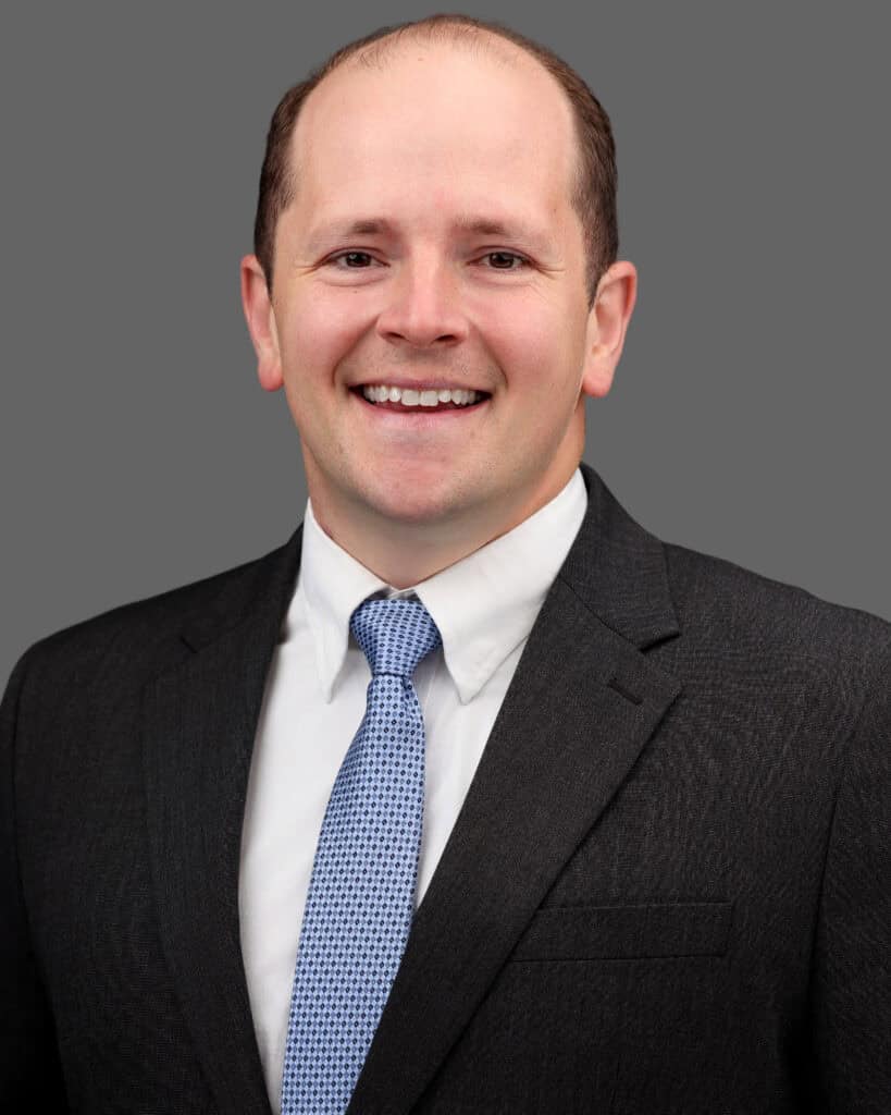 Professional headshot of a smiling man in formal attire against a gray background.